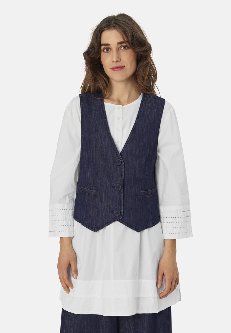 Dunkelblaue Jeansweste mit V-Ausschnitt, drei Knöpfen und zwei Fronttaschen, getragen über einem weißen Hemd mit strukturierten Ärmeln.