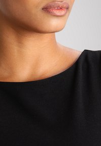 Närbild av en persons nedre ansikte, nacke och axel som bär en svart topp mot en enfärgad grå bakgrund.