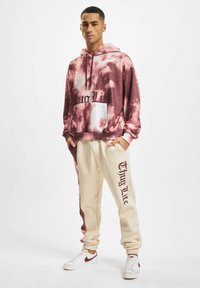 Felpa con cappuccio in sfumature di rosa, con tasca frontale, abbinata a pantaloni jogger crema con stampa "Thug Life" in bordeaux. Sneakers bianche.