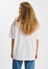 Biała, bawełniana t-shirt w oversize'owym rozmiarze z krótkim rękawem, charakteryzująca się luźnym krojem i gładką teksturą. Noszona z niebieskimi dżinsami.