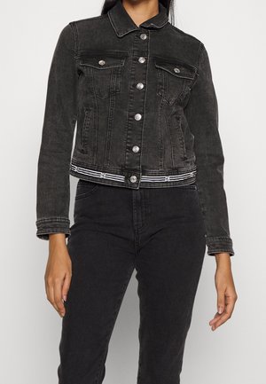 Kvinde iført en tætsiddende sort denimjakke med sølvknapper og sorte højtaljede jeans mod en ensfarvet baggrund.