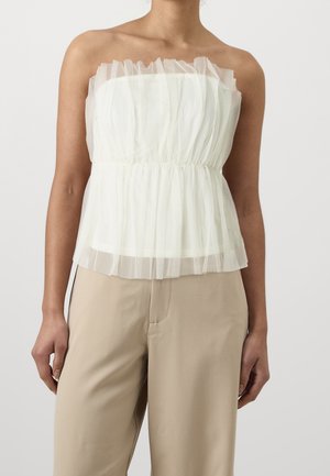 Kobieta w białym, bezramiączkowym, falbanianym przezroczystym topie i beżowych spodniach, stojąca na jednolitym jasnoszarym tle.