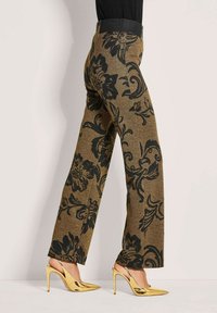 Bruine broek met zwart bloemenpatroon, gedragen met glanzende, gouden, hoge hakken slingback schoenen door een persoon die tegen een effen achtergrond staat.