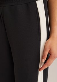 Gros plan sur une main reposant sur un pantalon noir avec une fine bande verticale blanche et une couture de poche latérale visible.
