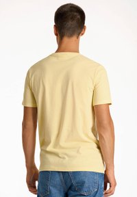 T-shirt di cotone gialla con colletto rotondo e maniche corte, texture liscia, design attillato, mostrata da dietro. Indossata con jeans in denim blu.
