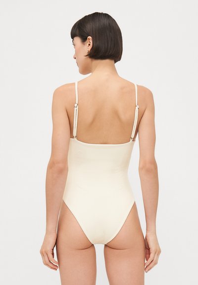 Einteiliger Badeanzug in Creme mit gerippter Textur. Verfügt über dünne, verstellbare Träger mit Beschlägen und ein hochgeschnittenes Bein-Design. Der Rücken ist vollständig entblößt.