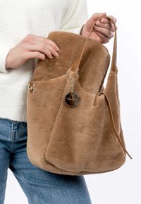 Sac seau en fausse fourrure beige avec une texture lisse, une sangle en cuir et une étiquette en métal. Présente une forme décontractée et un ouvert sur le dessus pour un accès facile.