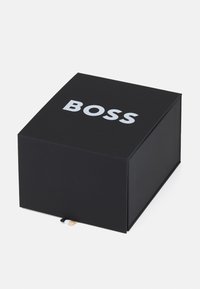 Boîte en carton noire avec une finition mate, présentant un large texte blanc "BOSS" sur le devant et une petite étiquette en tissu au bord inférieur.
