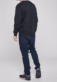 Svart bomullströja med lång ärm, ribbade muddar och nederkant; matchad med mörkblå jeans och grå sportskor med röda accenter.