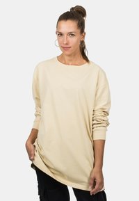 Beige langärmliges Oversized-T-Shirt mit Rundhalsausschnitt, mit herunterhängenden Schultern und gerippten Bündchen. Aus weichem Baumwollmaterial.