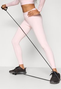 Haut court à manches longues rose clair et leggings taille haute assortis, associés à des baskets noires, tenant une bande de résistance pour exercice.