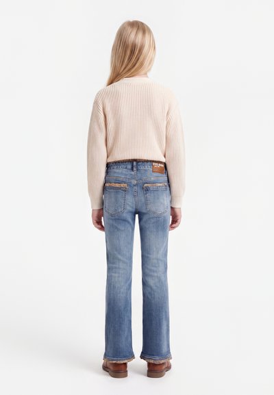 Enfant aux longs cheveux blonds, de dos, portant un pull en tricot crème, un jean bleu avec poches arrière et des chaussures marron sur fond blanc.