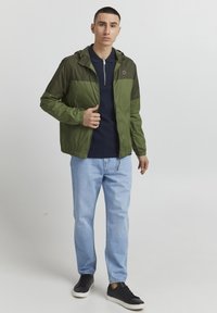 Olive green lightweight jacket with a hood, featuring a zip closure and elastic cuffs, worn over a navy polo and light blue jeans.