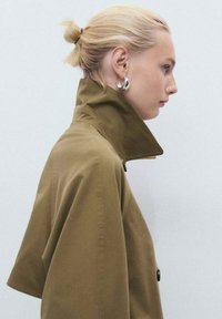 Un manteau marron à col haut, avec une texture lisse et des accents de boutons, porté par une personne avec une coiffure lisse et tirée vers l'arrière.