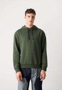 Felpa con cappuccio di colore verde scuro, realizzata in morbido tessuto. Presenta un cappuccio con cordino e polsini a costine, abbinata a pantaloni in denim a toppe.