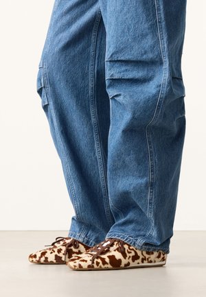 Benen met losse blauwe spijkerbroeken en platte veterschoenen met een bruin-wit koeienprintpatroon op een lichte vloer.