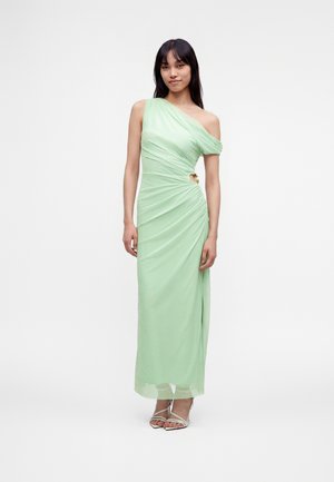 Femme debout dans une robe longue vert pâle à une épaule froncée avec une découpe sur le côté et portant des talons blancs à brides.