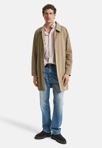 Beige lange jas met een kraag, gedragen over een gestreept shirt, gecombineerd met lichtblauwe jeans en zwarte schoenen. Soepele textuur, casual ontwerp.