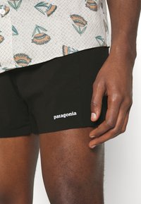 Black Patagonia swim shorts feature a snug fit, smooth texture, and white logo on the lower left side, paired with a patterned shirt.