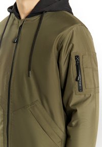 Giubbotto bomber verde oliva con cappuccio nero, tasche laterali e tasca con zip su un braccio, caratterizzato da una texture liscia e opaca e un design minimalista.