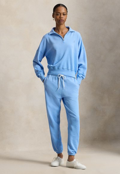 Γυναίκα που φορά ανοιχτό μπλε φούτερ με μισό φερμουάρ και ταιριαστό παντελόνι φόρμας με λευκά αθλητικά παπούτσια, στέκεται μπροστά σε ουδέτερο φόντο.