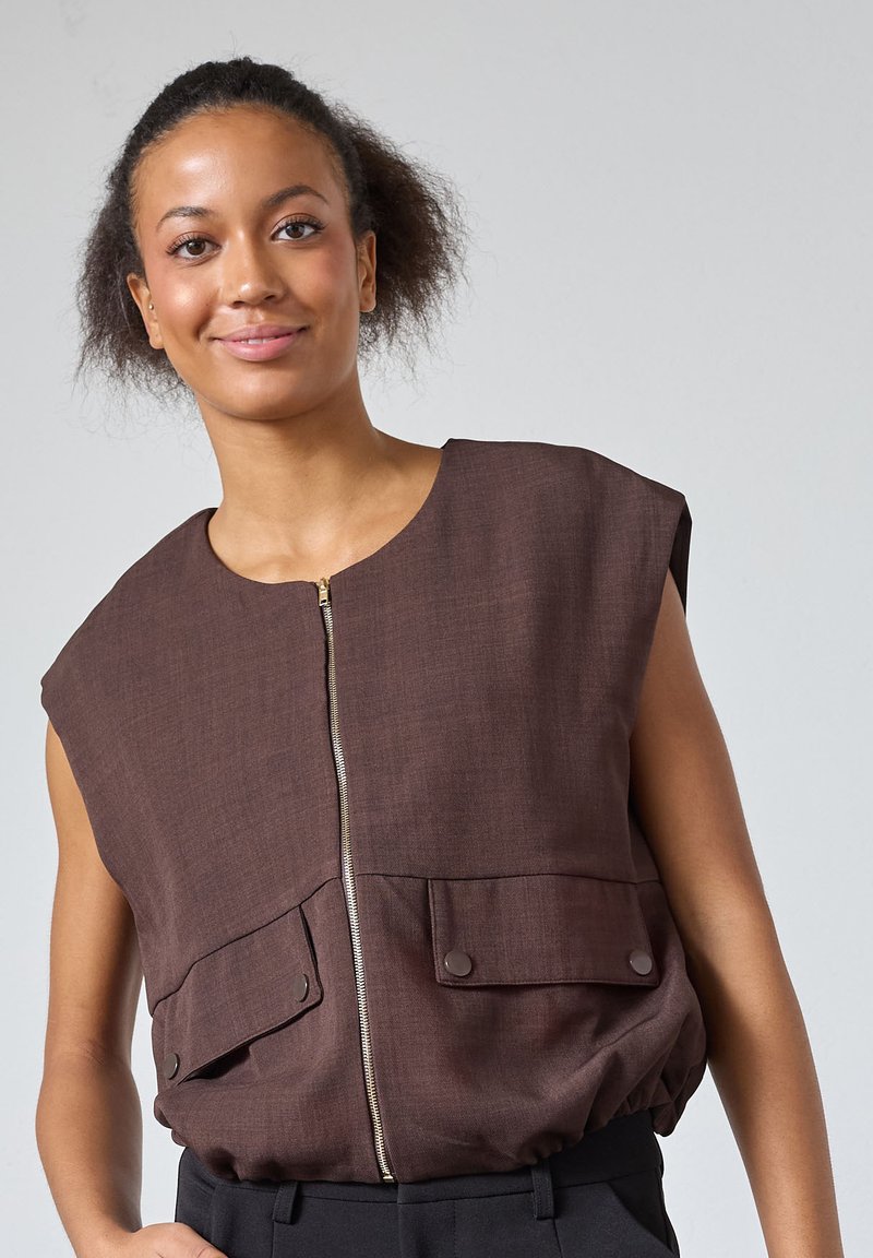 Brun ærmeløs top med rund halsudskæring, frontlynlås og to påsyede lommer. Stoffet har en struktureret finish med samlet afslutning ved kanten.