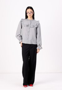 Grå blus med knäppning och volangkrage samt långa ärmar, med en texturerad mönstring. Kombinerad med svarta vida byxor och röda skor.