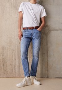 Una persona está de pie contra una pared de hormigón, vistiendo una camiseta blanca con texto, jeans azules y zapatillas altas blancas.