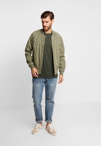 Jachetă bomber verde măslin, din material ușor, cu manșete și guler elastic. Asortată cu un tricou verde închis, blugi albaștri deschis și adidași bej.