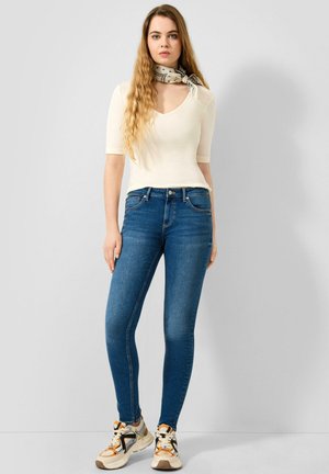 Jonge vrouw met lang golvend haar, draagt een wit topje met ronde hals, blauwe skinny jeans, sneakers met oranje accenten en een geprinte halsdoek.