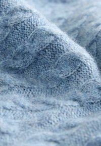 Lyseblå strikket stof med et struktureret kabelmønster og blød, fuzzy overflade. Nærbillede viser indviklede syninger.