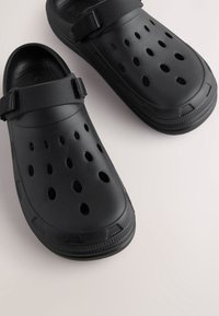 Sabots noirs en matériau synthétique avec plusieurs trous de ventilation, une bride texturée et une semelle robuste pour une meilleure adhérence et stabilité.