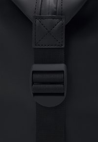 Spallaccio di zaino in nylon nero con una texture liscia, cinturino regolabile e una fibbia rettangolare con un motivo a punto croce.