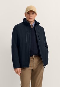 Marineblaue wasserdichte Jacke mit Frontreißverschluss, zwei Brusttaschen mit Reißverschluss und hohem Kragen, getragen über einem marineblauen Pullover und beigen Hosen. Beige Kappe inklusive.