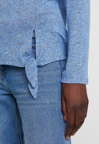 Ljusblå, texturerad långärmad topp med en knuten detalj vid fållen och subtila sidofickor, tillsammans med jeans.