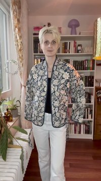 Pessoa com cabelo loiro curto, vestindo um casaco escuro com padrão floral e calças brancas, em pé numa sala com estantes de livros e plantas.