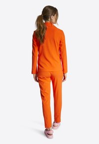 Oranje tweedelige outfit met een colbert top en slimfit broek. Textuurstof en schoenen in neutrale tinten maken de look compleet.