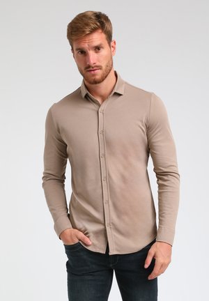 Camisa de botones de manga larga beige con una textura suave, corte ajustado y dobladillo curvado, combinada con jeans de mezclilla oscura.