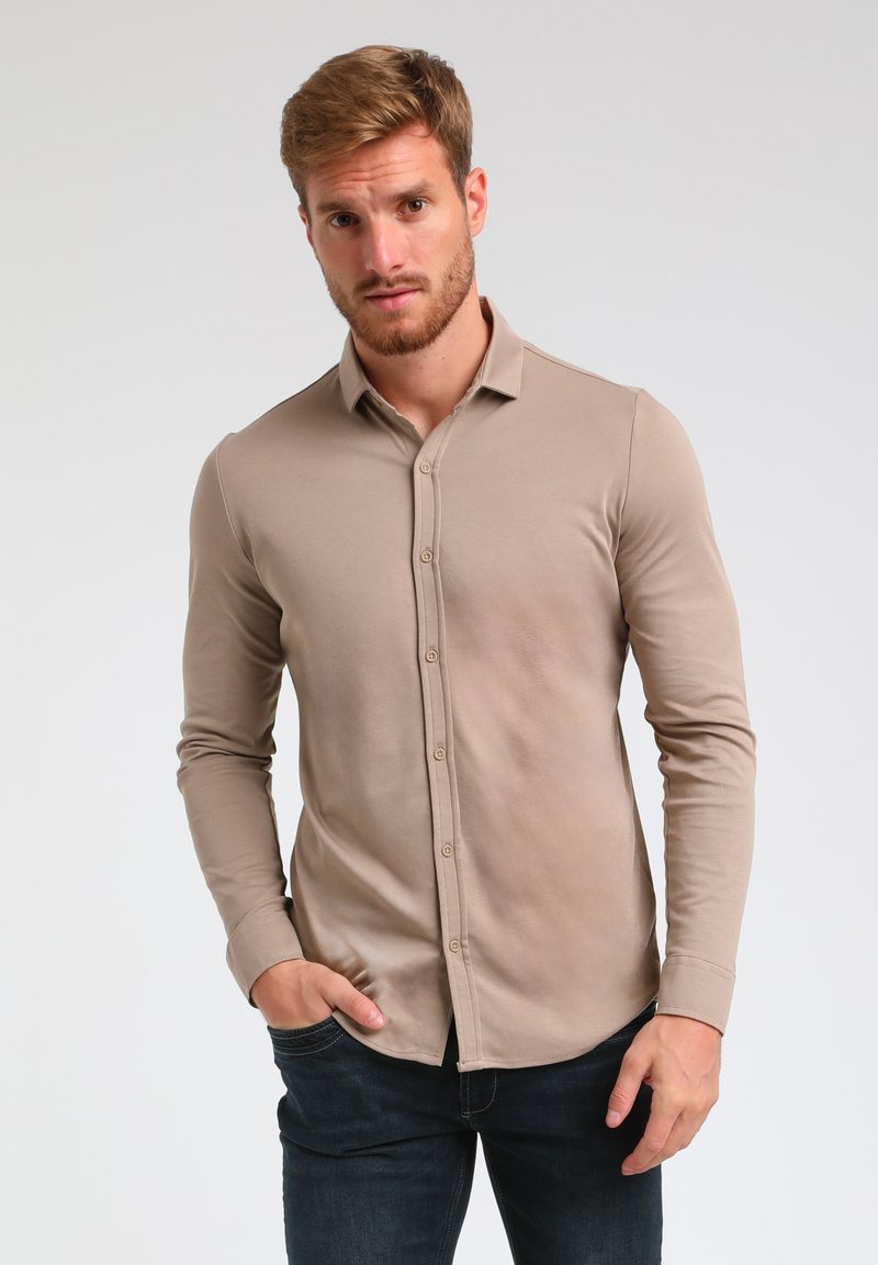 Beige Langarmhemd mit Knopfleiste und weicher Textur, schlanker Schnitt und abgerundetem Saum, kombiniert mit dunklen Jeans.