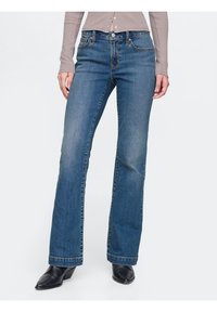 Kvinna klädd i blå raka jeans, ljusbrun långärmad topp och svarta spetsiga klackstövlar, stående mot vit bakgrund.