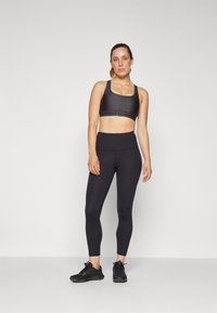 Sostenedor deportivo negro con tirantes anchos y banda inferior con logotipo, combinado con leggings negros de cintura alta. Zapatillas deportivas negras completan el conjunto.