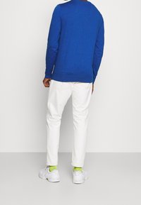 Blauer gestrickter Pullover mit langen Ärmeln und Rundhalsausschnitt. Kombiniert mit weißen Hosen und weißen Turnschuhen, die helle grüne Socken tragen.