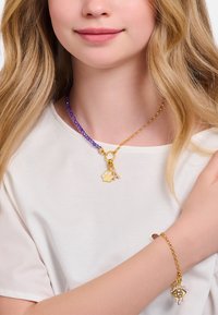 Lila Perlenkette mit goldfarbenen Beschlägen und zwei Anhängern; einer ist ein Pfotenabdruck, der andere zeigt ein funkelndes Auge. Goldene Gliederarmband eingeschlossen.