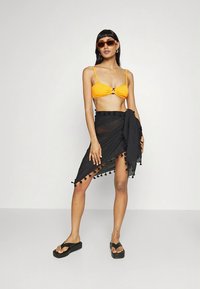 Oranžen bikini zgornji del s središčnim obročkom, kombiniran z črno prozorno krilno ovojnico z obrobjem iz cofkov in črnimi platformnimi sandali.