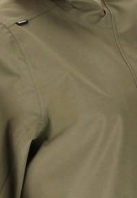 Lähikuva oliivinvihreän takin kankaasta, jossa näkyy saumaviivat ja pieni musta vetoketjun merkki olkapääalueella.