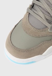 Demonstrativo em close-up de um bico de ténis taupe com painéis de camurça e couro, malha cinza, atacadores grossos, entressola branca e detalhe da sola azul clara.