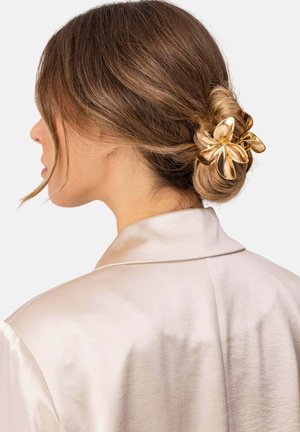 Frau mit hellbraunen Haaren, zu einem niedrigen Dutt gestylt und mit einer großen glänzenden goldenen Blumen-Haarklammer befestigt, trägt ein seidiges beiges Oberteil.