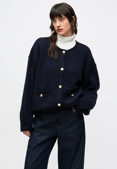 Cárdigan de punto azul marino con botones dorados, dos bolsillos delanteros, corte relajado y puños acanalados. Combinado con pantalones de mezclilla oscura.