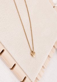 Colier cu pandantiv în formă de inimă din aur, pe un lanț delicat, expus pe o țesătură bej moale cu suprafață texturată și margine din lemn.