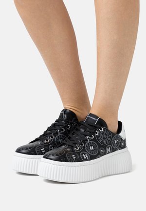 Jambes portant des baskets plateformes noires avec des motifs circulaires blancs de logo et des semelles blanches épaisses et striées, photographiées sur un fond clair uni.